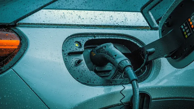 Elektroauto uzlādes procesa tuvplāns ar kabeli pieslēgtu pie automašīnas lietainā laikā.