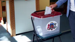 Vēlētājs ievieto vēlēšanu aploksni caurspīdīgā plastmasas vēlēšanu urnā ar Latvijas valsts ģerboņa uzlīmi.