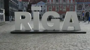 Liels, balts uzraksts RĪGA uz bruģētas vecpilsētas laukuma fona Rīgā.
