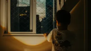 Mazs bērns vakarā raugās pa logu uz pilsētas gaismām, simbolizējot bērnības atmiņas.