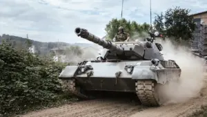 Militārais tanks pārvietojas pa zemes ceļu Austrumu pierobežas aizsardzības izbūves darbu kontekstā.