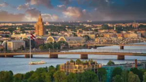 Plašs skats uz Rīgas panorāmu ar Latvijas karogu un Daugavu saulrieta gaismā.