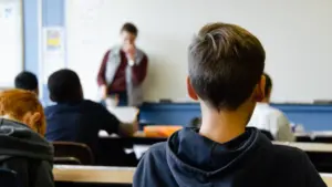 Skolēni klases telpā vēro skolotāju, kurš stāv pie baltās tāfeles.